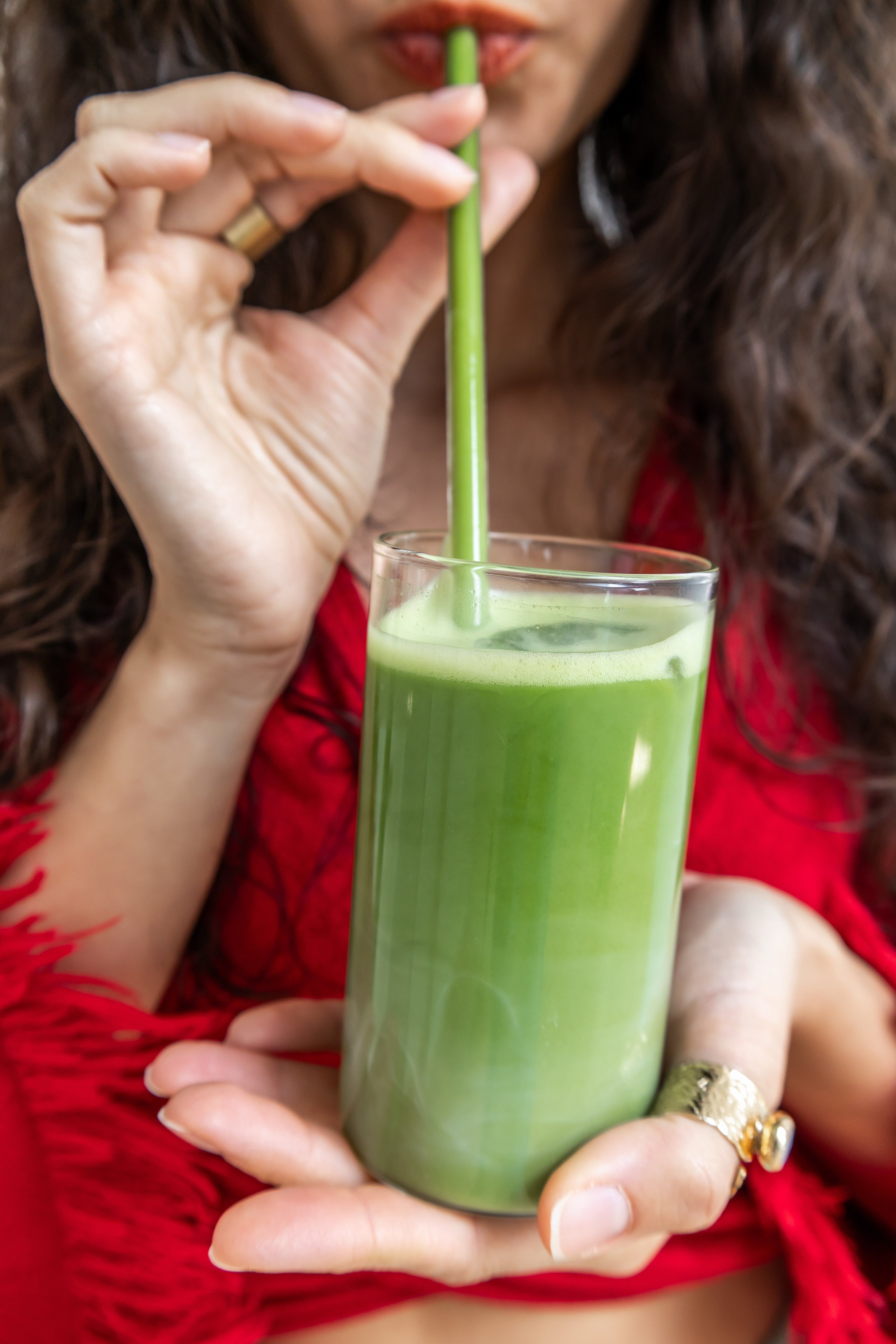 Person drinking a matcha latte from a glass with a straw, wearing a red garment.