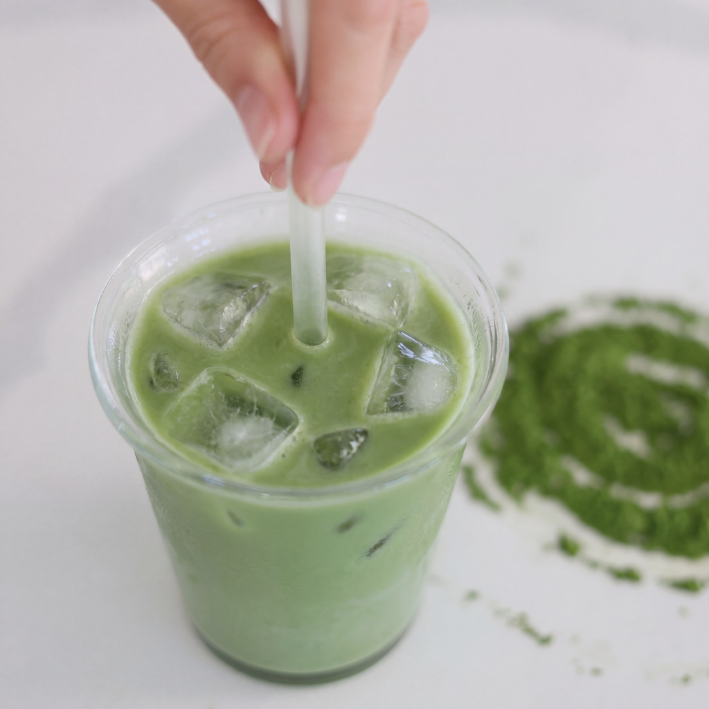 Hand stirring an iced matcha latte with a clear straw in a glass cup, with matcha powder scattered on a white surface.