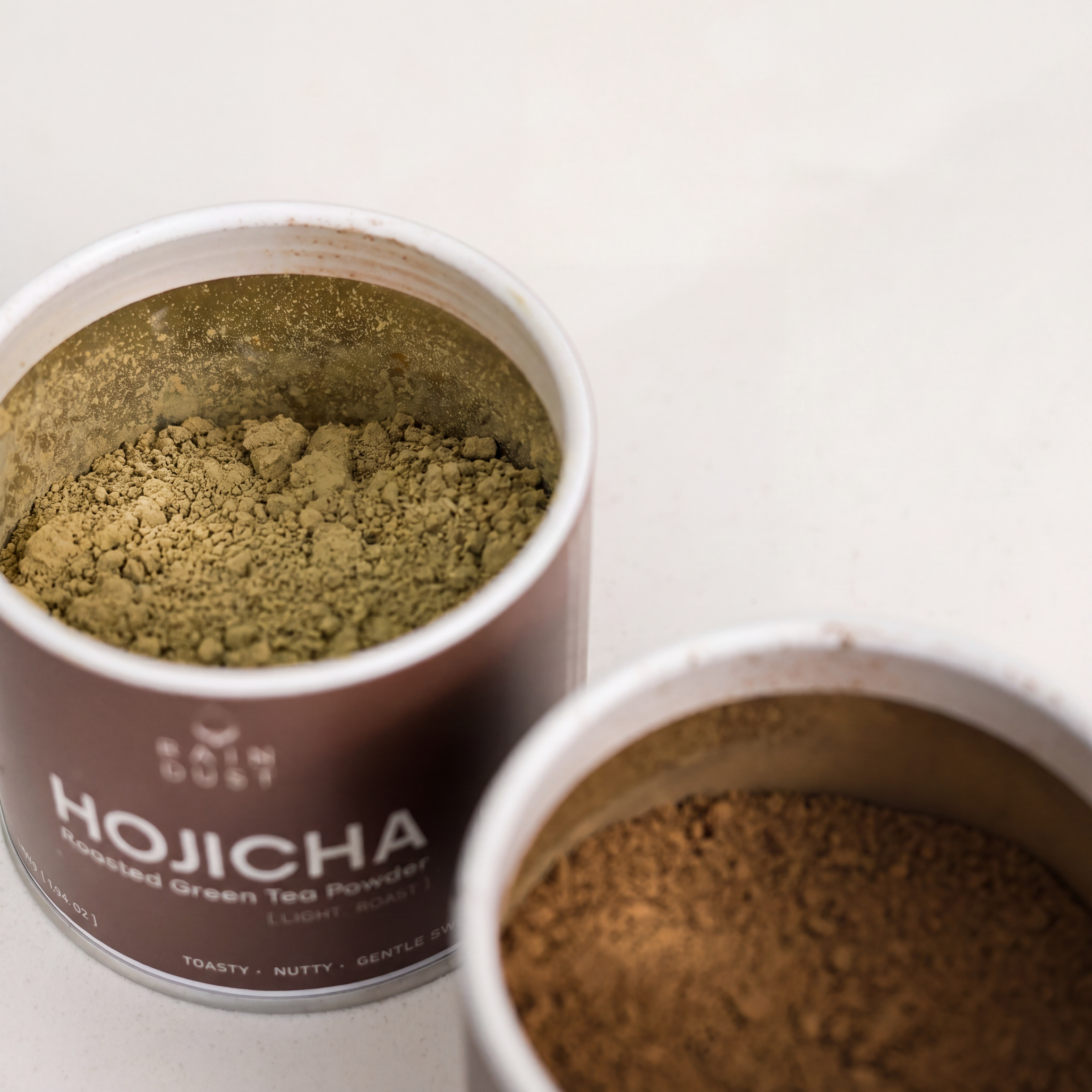 Two open tins of Rain Dust Light Roast Hojicha powder showing finely milled Japanese roasted green tea on a neutral background