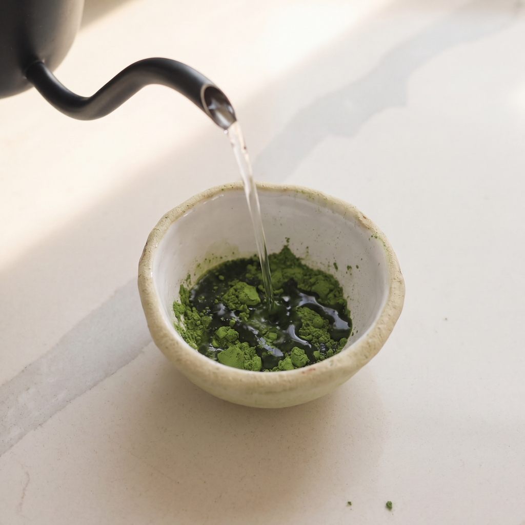 Ceremonial grade Japanese matcha powder in a small ceramic bowl with hot water being poured from a black kettle on a light beige surface.